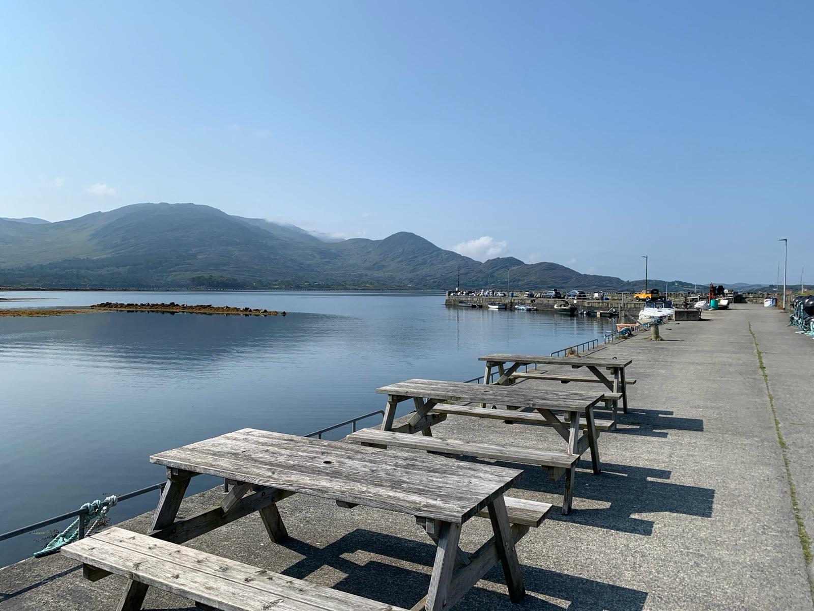 A quiet Irish harbour pier with wooden picnic benches, boats moored, and mountains beyond