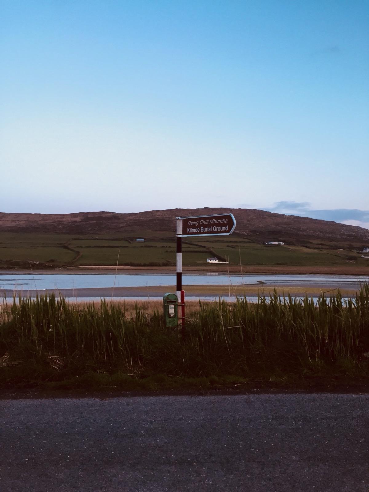 A bilingual Irish road sign by an estuary at dusk, pointing to Kilmoe Burial Ground